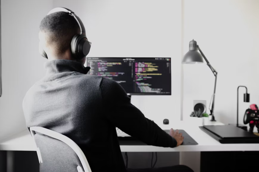 A male individual wearing headphones is seated at a desk, operating a computer.png